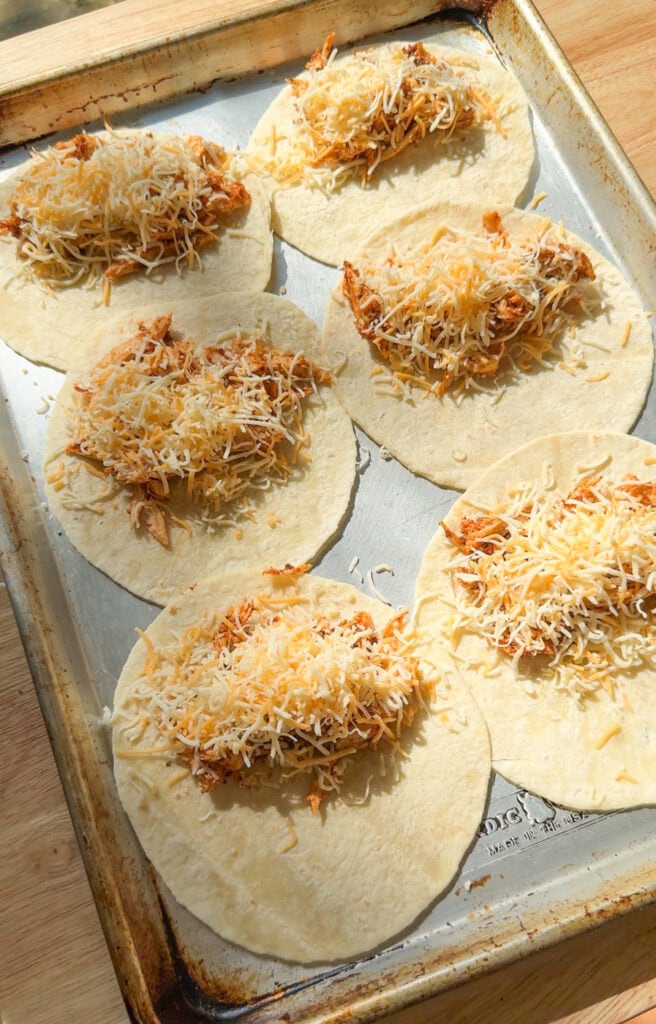 Shredded chicken and cheese added to tortillas on a sheet pan for baked chicken tacos