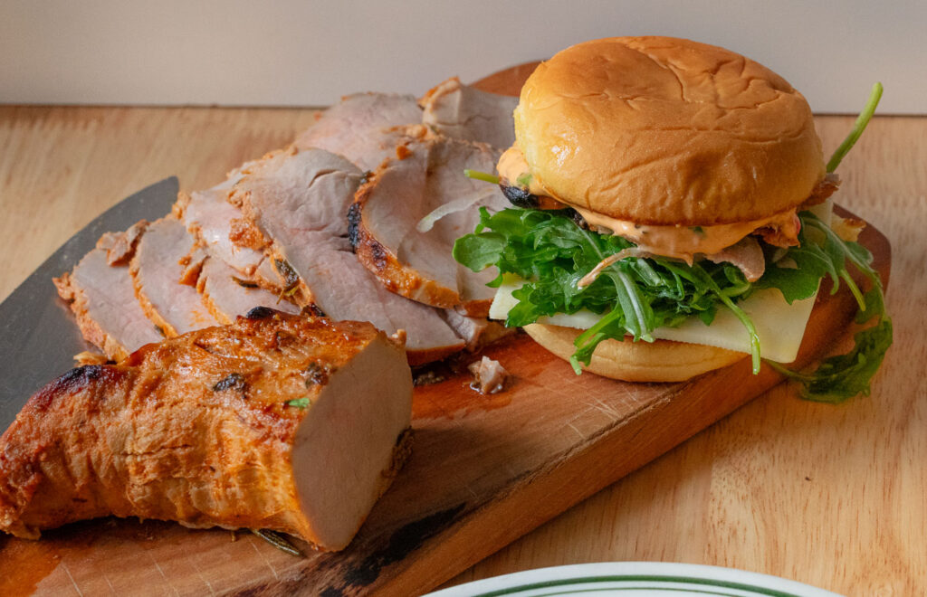 Sliced pork tenderloin on a wooden board next to a pork tenderloin sandwich.
