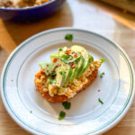 Spicy egg salad on crispy hash brown toast topped with sliced avocado and chili crisp on a white plate.