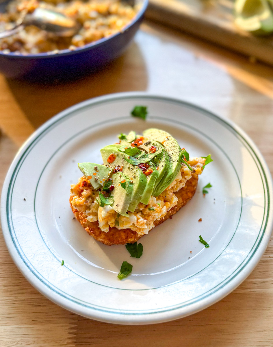 Spicy egg salad on crispy hash brown toast topped with sliced avocado and chili crisp on a white plate.