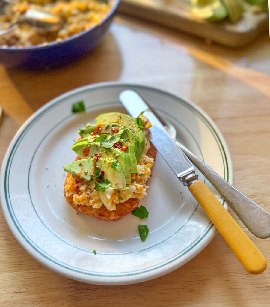 Hash brown toast topped with spicy egg salad, fresh avocado, herbs, and chili crisp.