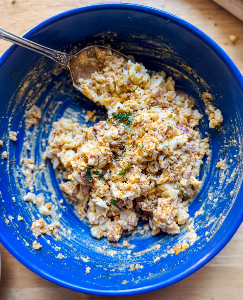 Spicy egg salad being mixed in a blue bowl with chili crisp and herbs.