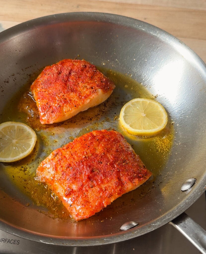 blackened cod cooking in skillet with lemon slices and butter