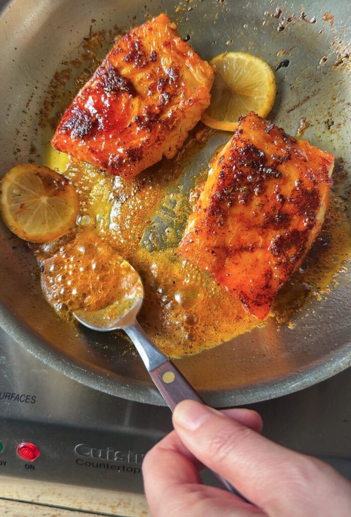 blackened cod in skillet with butter sauce and lemon while spooning sauce over fish