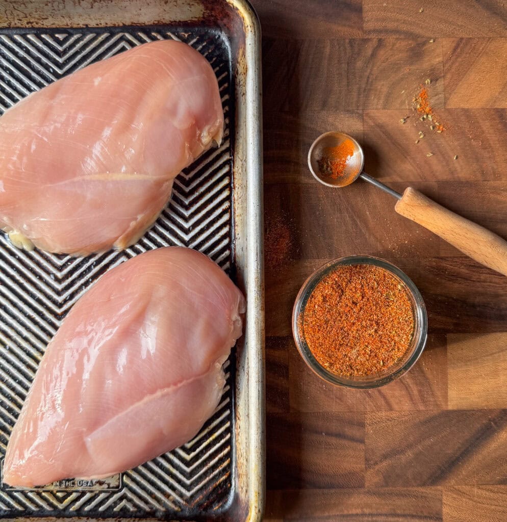 raw chicken breasts on a sheet pan with homemade chicken taco seasoning and a measuring spoon