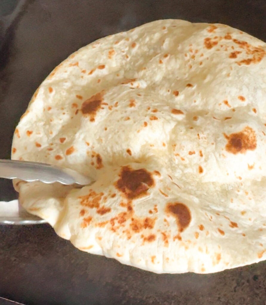 Flour tortilla puffing over open flame with tongs, lightly charred and blistered for grilled chicken wraps