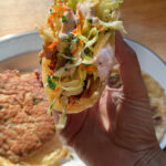 Hand holding a crispy Asian smash taco topped with crunchy cabbage slaw and spicy mayo.