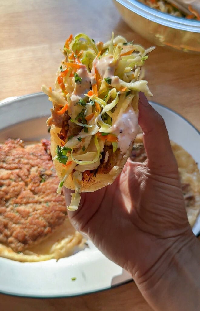 Hand holding a crispy Asian smash taco topped with crunchy cabbage slaw and spicy mayo.